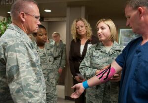 A physical therapy technician shows a brigadier-general and two other officers Kinesio Tape on a patient’s arm.